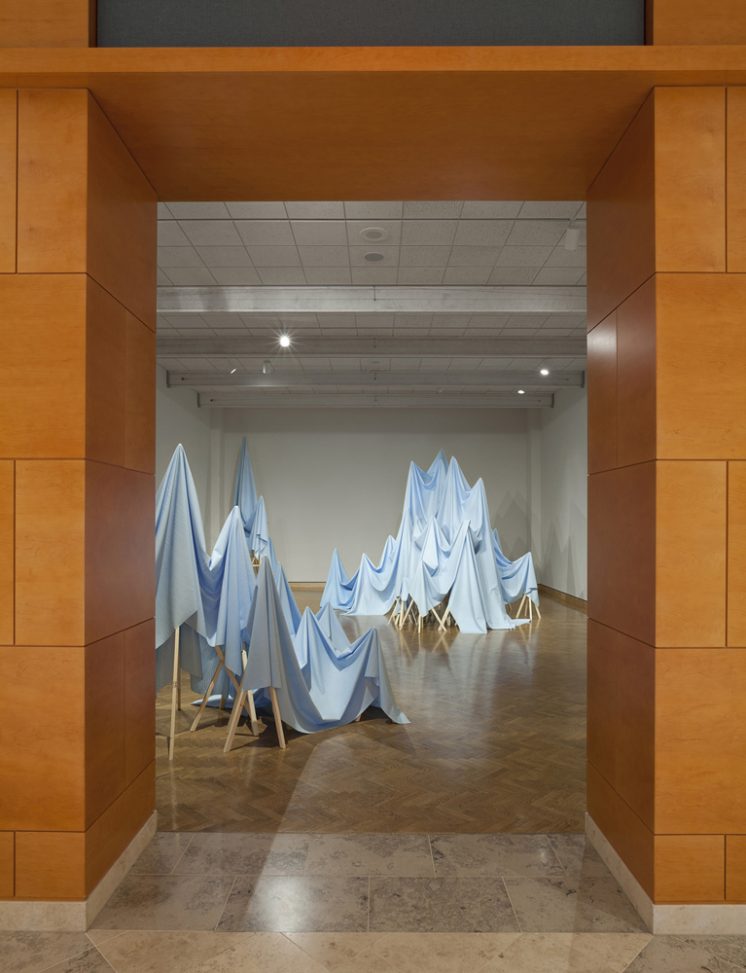 Posture is Everything, 2013. Sculptural installation: Silicone rubber, wood. 1352 square feet of sky blue rubber sheeting was produced to equal the dimensional area of this gallery space by painting layers of liquid silicone molding rubber onto a wall and then removing the cured material to create the objects within this gallery. The weighty rubber sheets are draped and given form over a series of wooden stands. The articulated material within the space was composed to reinterpret the physical gallery dimensions into an abstracted, undulating and peaked topography.